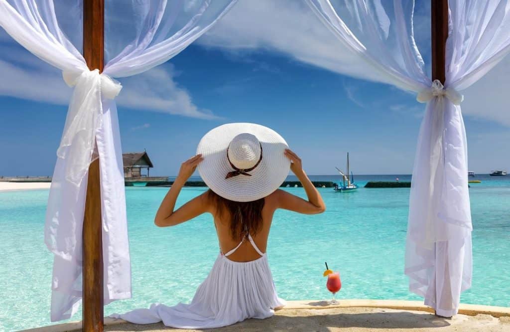 A woman sitting down and facing the ocean as she is wearing a white dress and a white beach hat.
