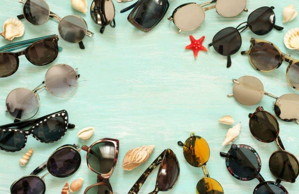 Several sunglasses arranged in a circle with a light blue background and seashells around them.