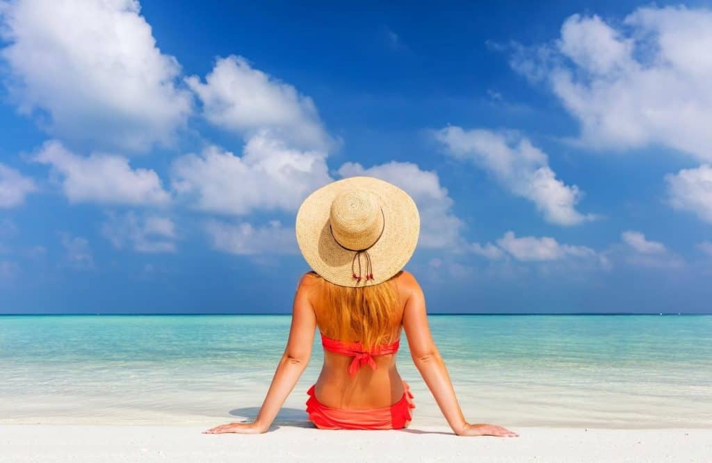 A woman in a red swimsuit with a brown sunhat that's essential on a Maldives packing list on sitting on the sand on the beach with blue skies