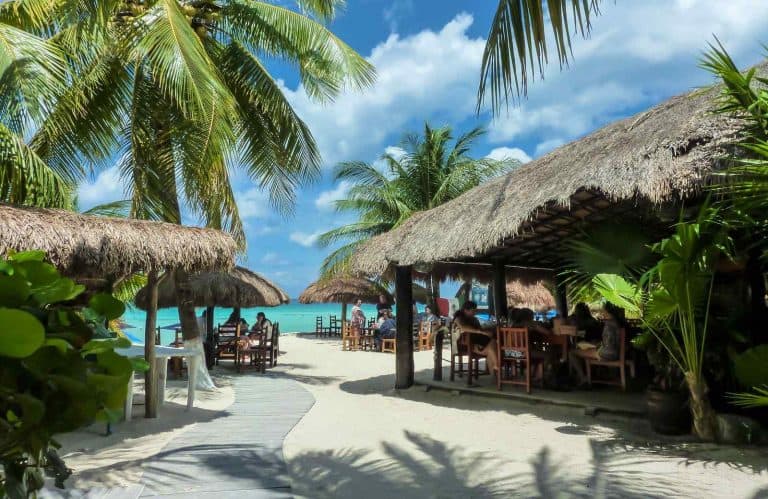 Two beach huts with palm trees above them and beautiful blue waters for is Cozumel safe.