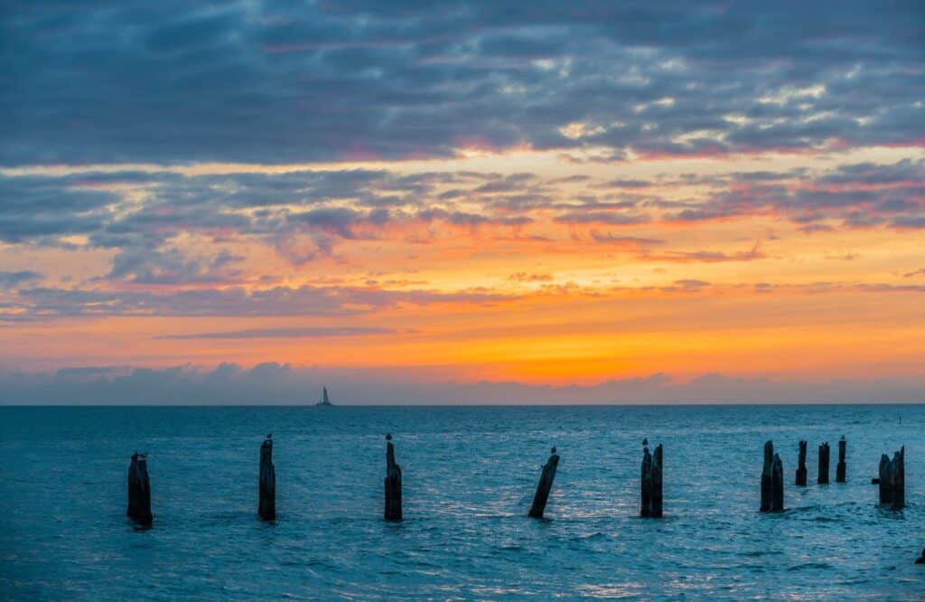 A sunset along the ocean with pastel orange and hue colors with a sailboat in the background, which the views make for the best sunset cruises in Key West.