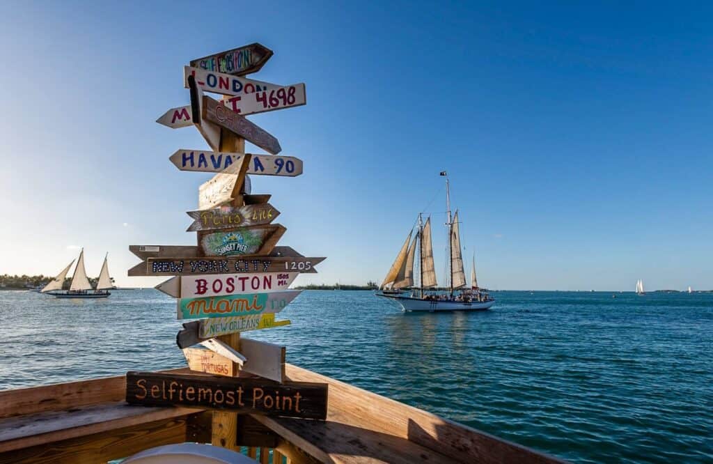 Two sailboats on the ocean going on a sunset sail in Key West with a wooden sign of cities and their distance from Key West.