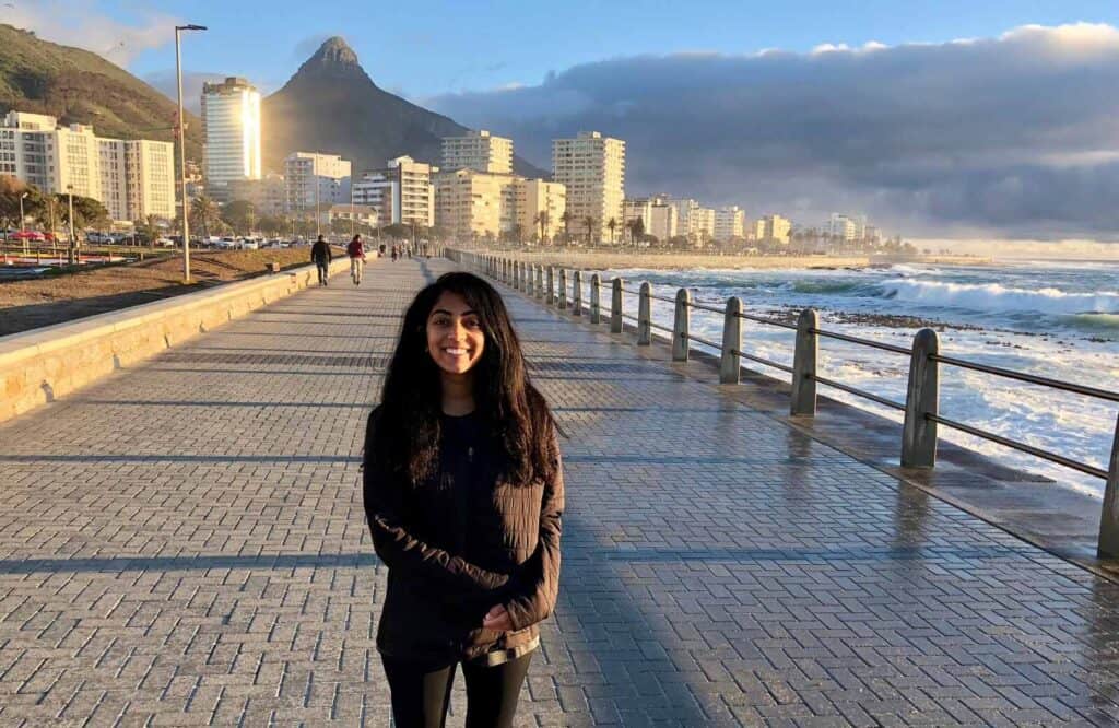 A woman standing on a pathway that runs along the ocean with a mountain in the background in Cape Town.