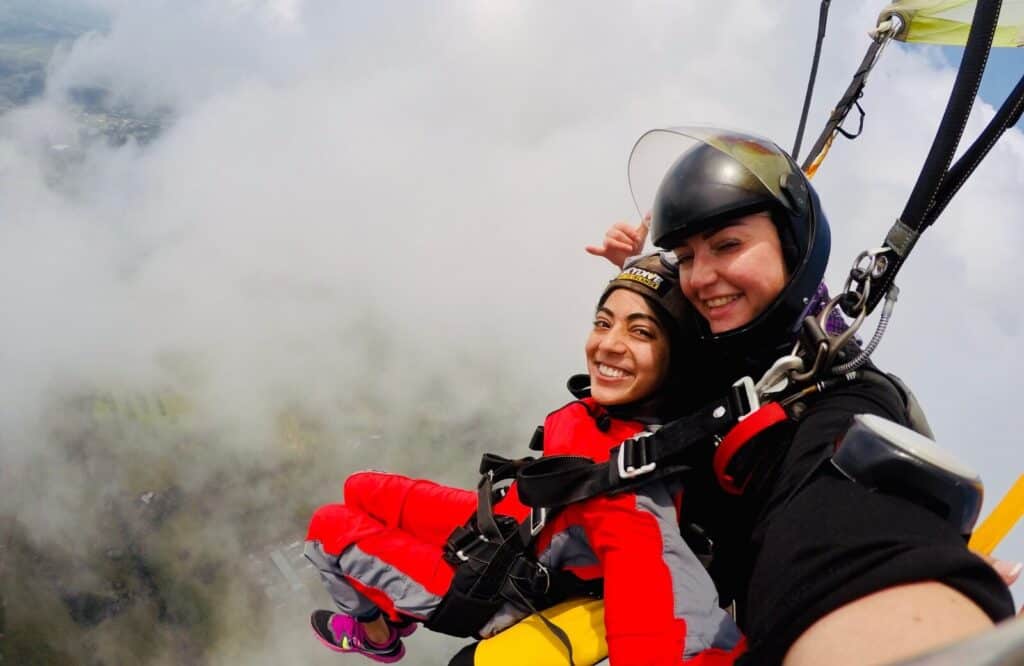 A woman in a red jumpsuit attached to another woman in a black jumpsuit while skydiving in New Zealand.