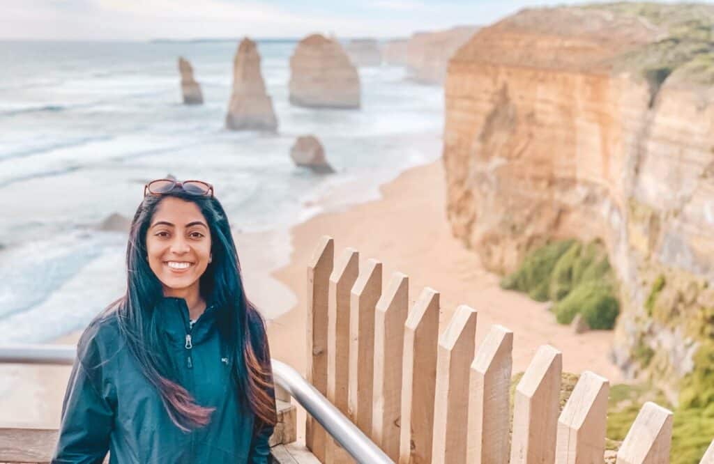 A woman standing in front of the Twelve Apostles in Australia, which are towering rocks emerging from the ocean.