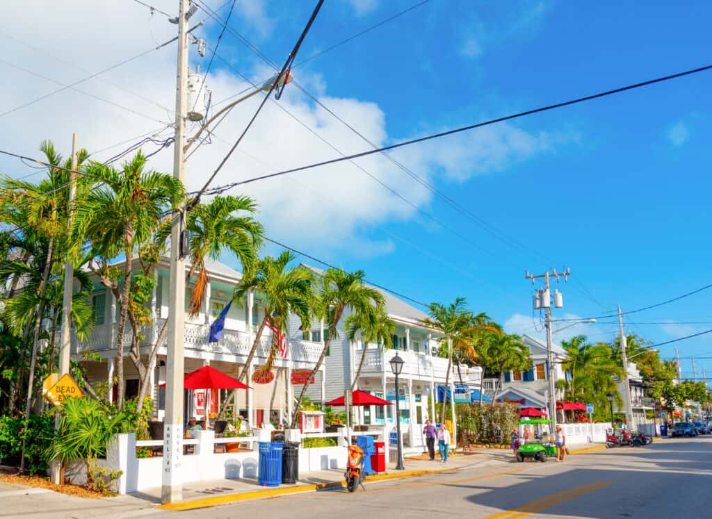 A sunny street scene in Key West with tropical palm trees, colorful umbrellas at an outdoor cafe, and traditional white-painted houses with verandas. Pedestrians walk along the sidewalk, and vehicles, including a scooter and a golf cart, are parked or in motion, under a bright blue sky scattered with clouds.