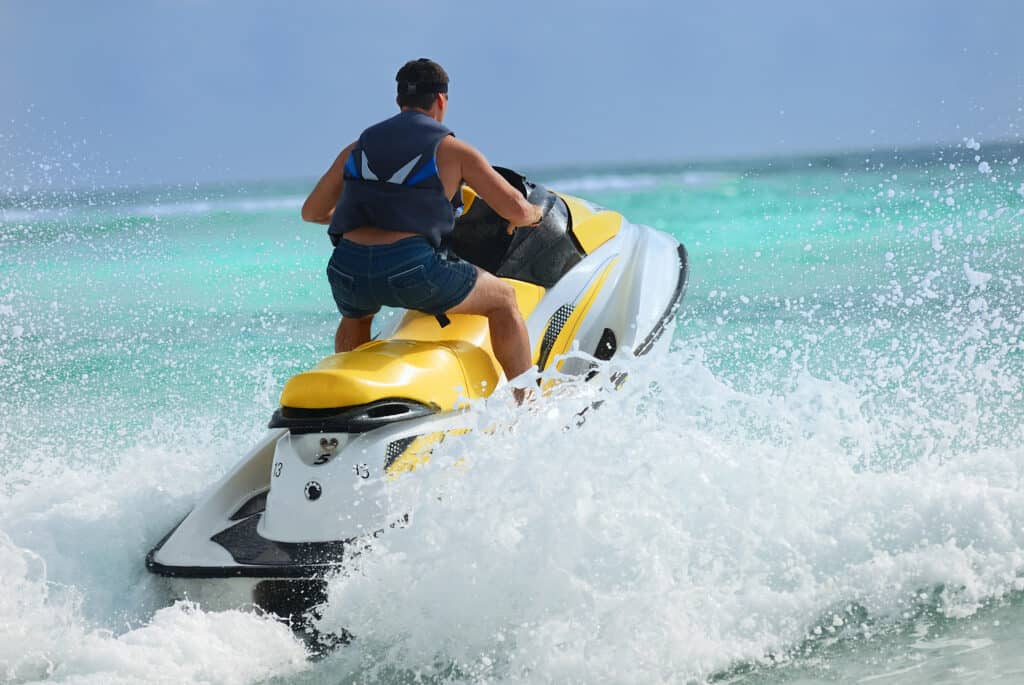 A person in a life vest is riding a yellow and white jet ski over turquoise waters, creating a splash in the sea. They appear focused as they maneuver the watercraft, with a clear sky and the horizon visible in the background.