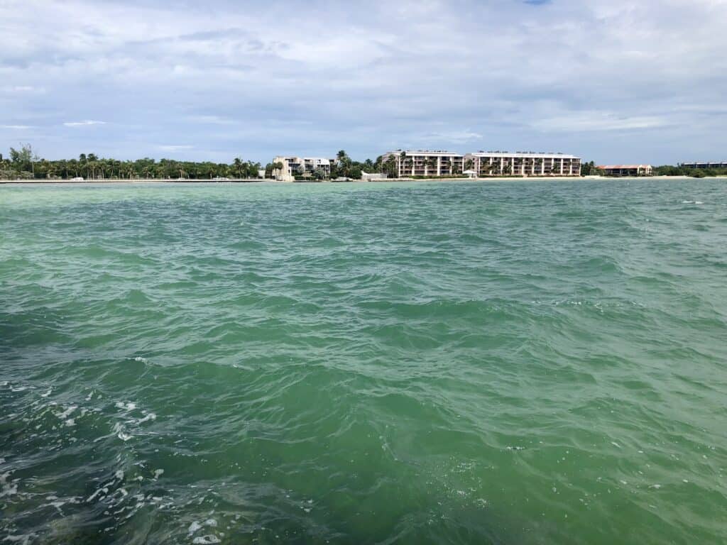 View of a calm turquoise sea with gentle waves, overlooking a coastline with lush greenery and partially constructed buildings in the background under a clear sky.