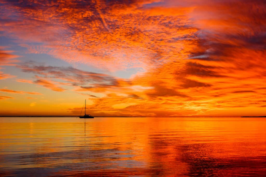 A serene sunset over calm waters with a sky painted in vibrant shades of orange, red, and yellow. A solitary sailboat is silhouetted against the fiery colors reflected on the water's surface, suggesting a peaceful evening at sea.