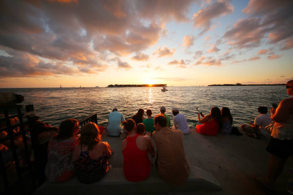A group of people sit along the waterfront, enjoying a picturesque sunset with warm hues reflecting off the water. Silhouettes of sailboats are visible against the glowing horizon as the sun dips low, casting a tranquil scene over the gathering.