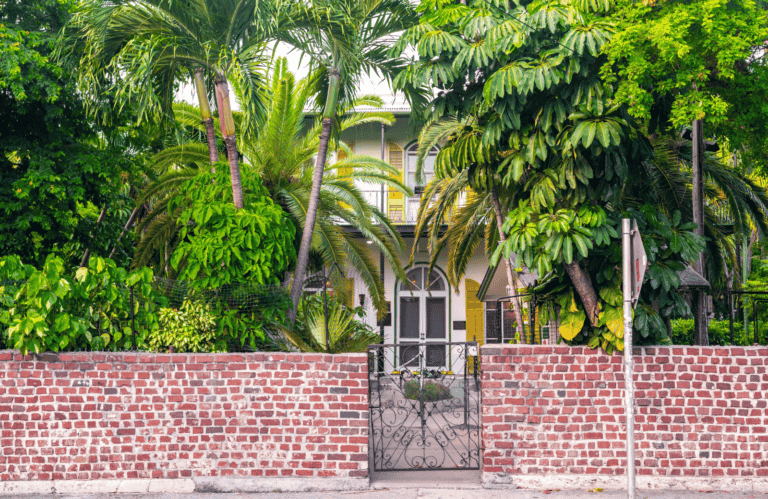 A charming, historic house in Key West is partially obscured by lush tropical plants and trees. The entrance features a wrought iron gate set into a red brick wall, leading to a path flanked by greenery. The house has a welcoming facade with yellow shutters and a prominent arched doorway.