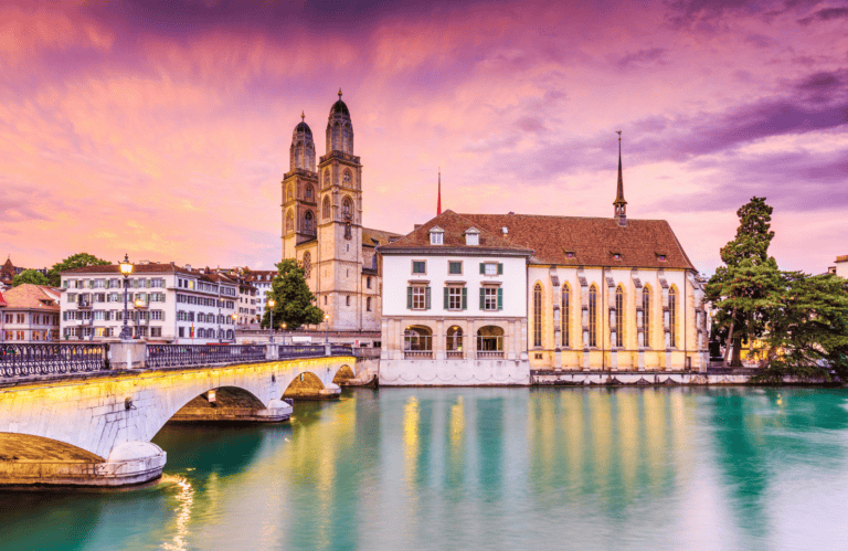 The image depicts a scenic view of Zurich, Switzerland, showcasing the iconic Grossmünster church with its twin towers, a bridge over the Limmat River, and traditional buildings along the water. The sky is painted with vibrant hues of pink and purple, reflecting the setting sun. The calm water and historical architecture create a picturesque, serene atmosphere.