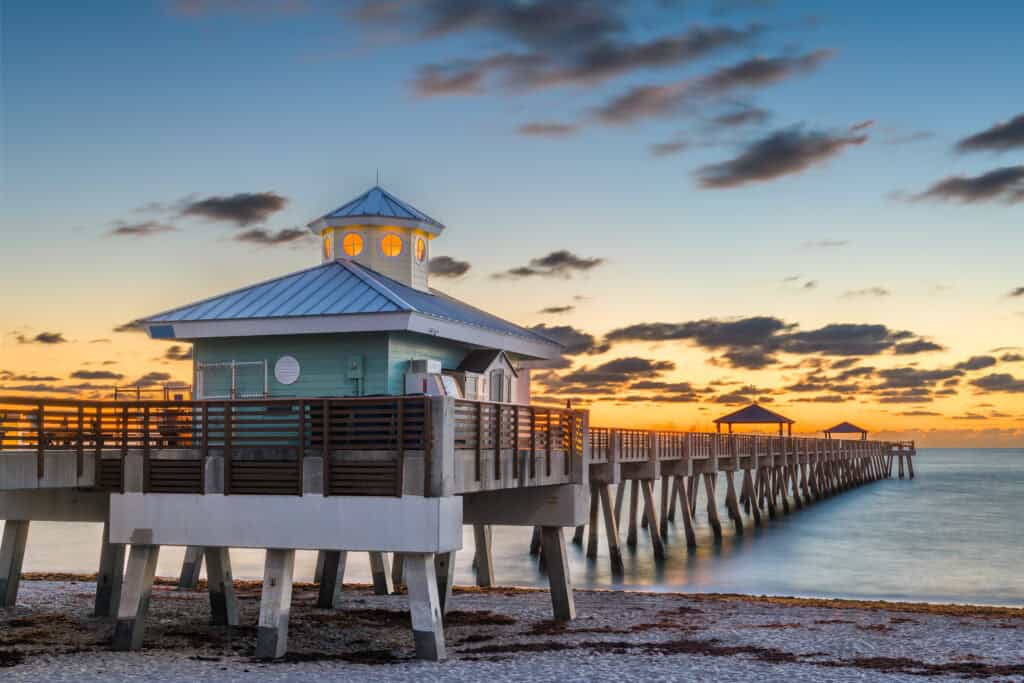Juno Beach Pier at sunrise, featuring a light blue pavilion on stilts with a metal roof and softly lit windows. The pier extends into the calm ocean, while the sky is illuminated with warm orange and yellow hues, creating a tranquil seaside scene.