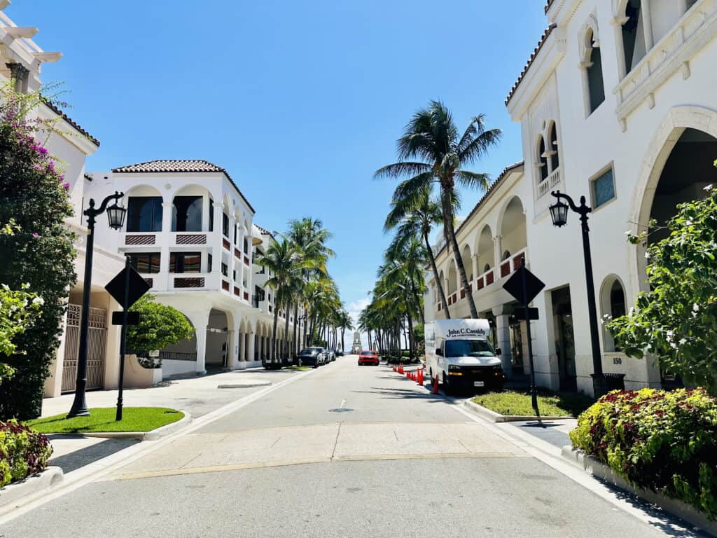 Street view in Palm Beach, Florida, featuring Mediterranean-style buildings with arched windows and white facades. The road is lined with palm trees and manicured greenery, creating a picturesque coastal town atmosphere under a clear blue sky.