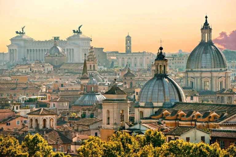 A panoramic view of Rome’s historic cityscape at sunset, featuring a cluster of domed churches and classical buildings with terracotta rooftops. The white marble Altare della Patria, with its iconic winged chariots, stands prominently in the background, bathed in warm golden light.